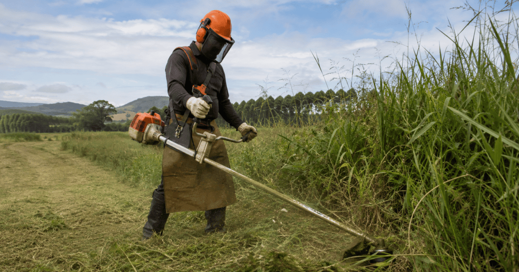Profissional utilizando roçadeira a gasolina em área rural com vegetação alta, usando capacete, protetor auricular, óculos de proteção, luvas e perneiras.
